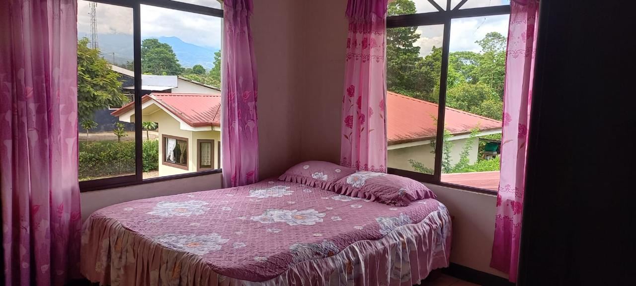 Bedroom with a pink floral bedspread and matching pillows, flanked by large windows and pink sheer curtains showing houses and trees outside.