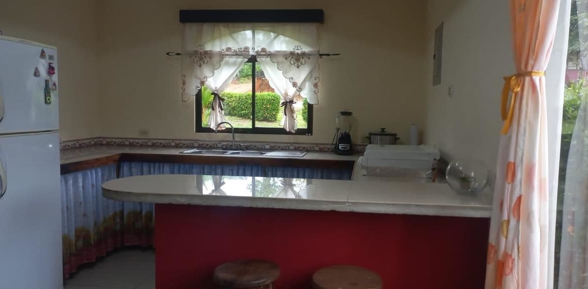 Kitchen with a white refrigerator, L-shaped counter, and central island under a window with lace curtains.