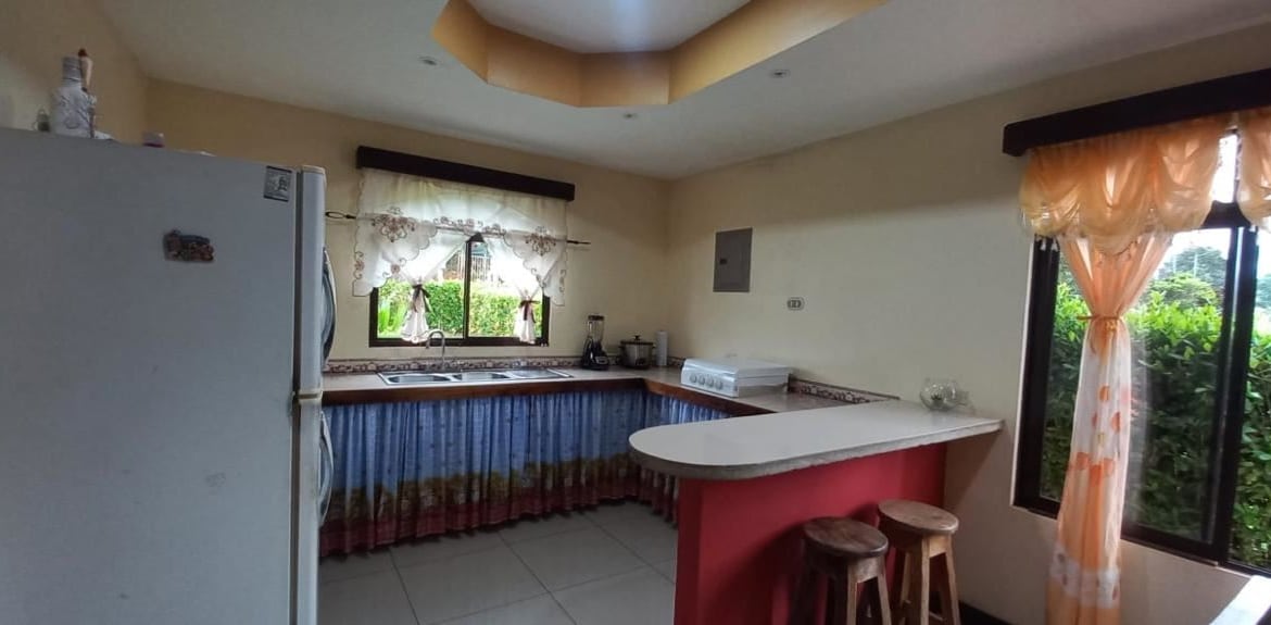 Kitchen with a white fridge on the left, L-shaped counter, and pink island bar with two wooden stools; window with lace curtains in the back and orange drapes by a large glass door to the right.