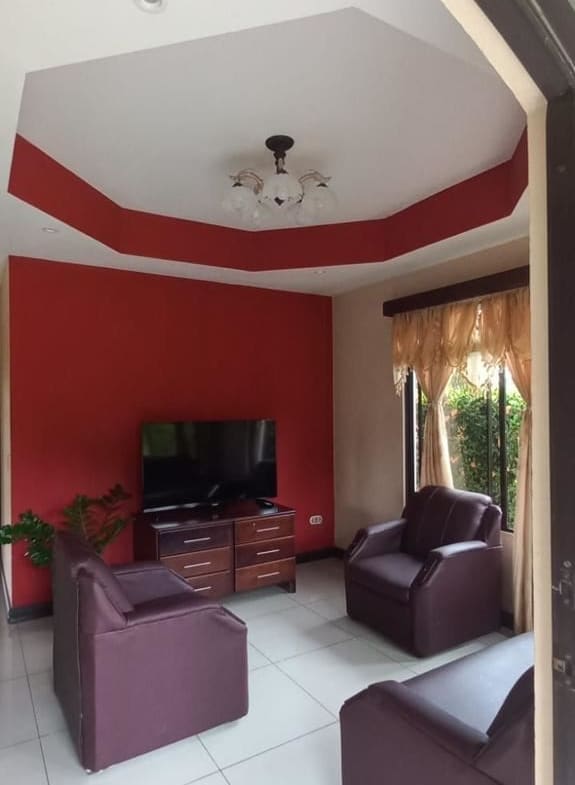 Living room with a red accent wall, four maroon leather chairs, and a TV on a wooden stand opposite the seating area, beige curtains by a window.