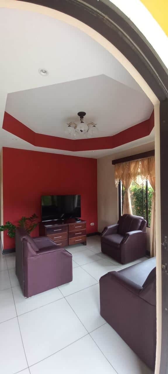 Living room with a red accent wall, four maroon leather chairs, and a TV on a wooden stand opposite the seating area, beige curtains by a window.