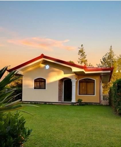 Single-story yellow house with a red roof and white columns, arched windows, on a green lawn at sunset.