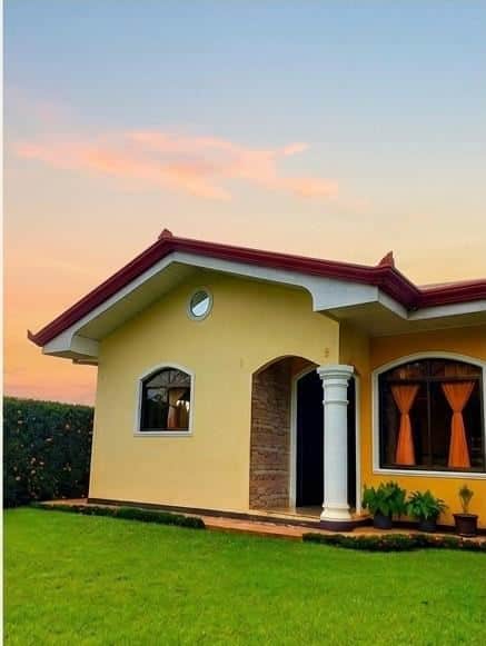 Yellow single-story house with red roof and white column porch, green lawn, and sunset sky behind it.