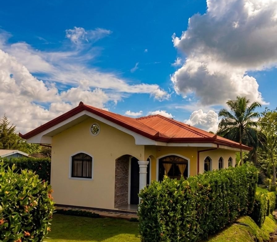 Single-story yellow house with a red roof, arched windows, and a manicured hedge in a tropical yard under a blue sky.