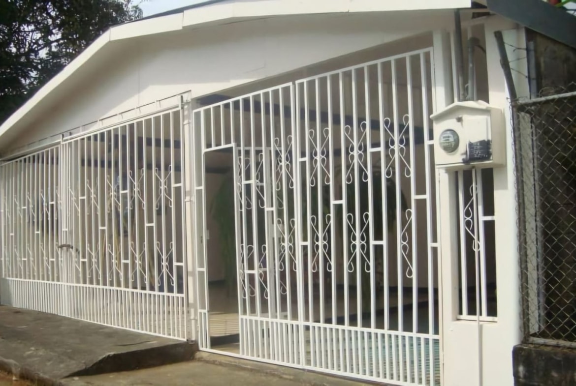 White decorative metal gate enclosing a small house, with a sloped roof and a mailbox on a pillar outside the gate