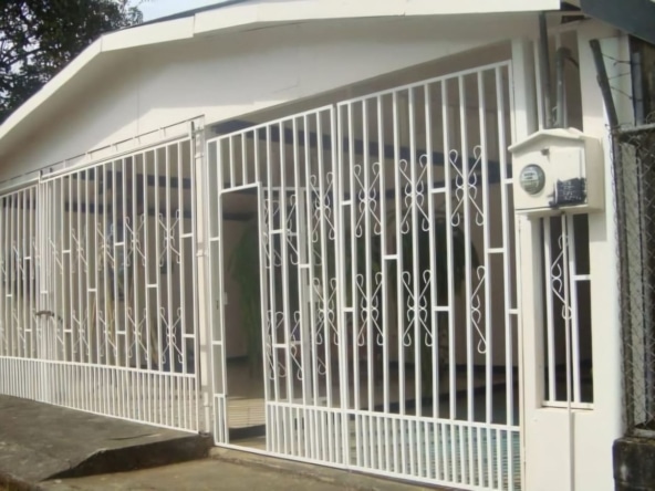 White decorative metal gate enclosing a small house, with a sloped roof and a mailbox on a pillar outside the gate