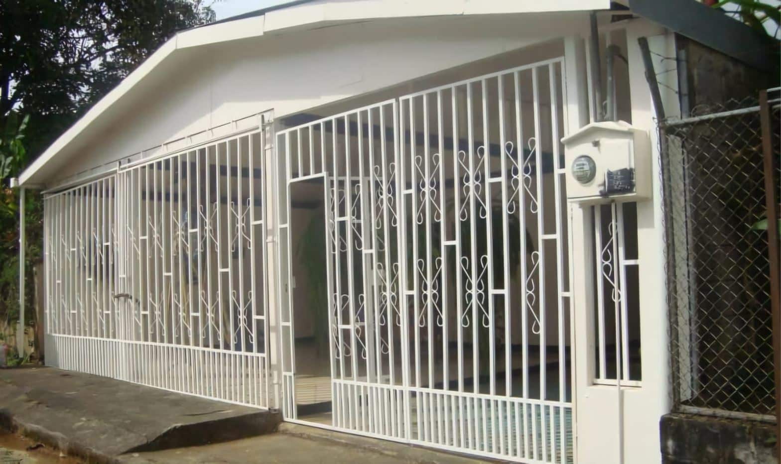 White decorative metal gate enclosing a small house, with a sloped roof and a mailbox on a pillar outside the gate