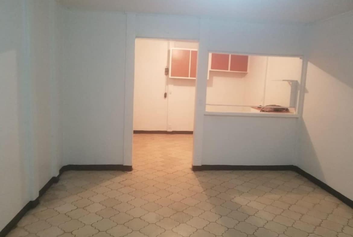 Empty basement-style room with pale blue walls, patterned tile floor, and a doorway opening to a bright kitchenette visible through a pass-through window