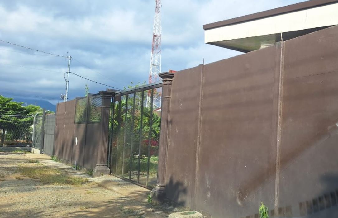 Tall brown boundary wall with a glass-paneled gate along a dirt path; cloudy sky and a communications tower in the background.