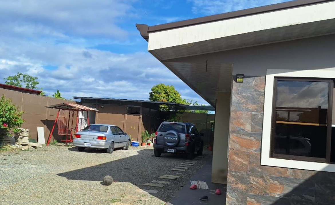 Modern house exterior with a gravel driveway and two parked cars (silver sedan and dark SUV) under a carport, stone-clad wall on the right.