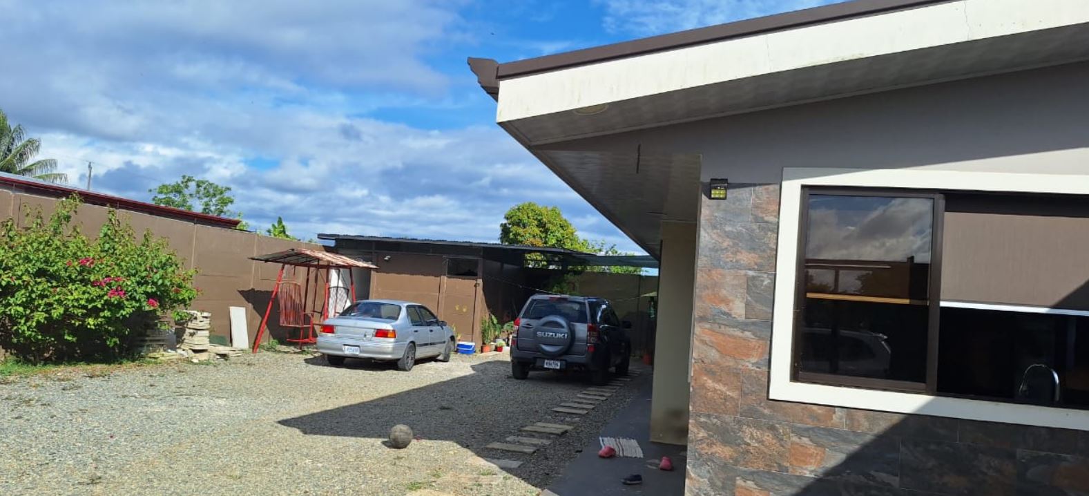 Modern house exterior with a gravel driveway and two parked cars (silver sedan and dark SUV) under a carport, stone-clad wall on the right.