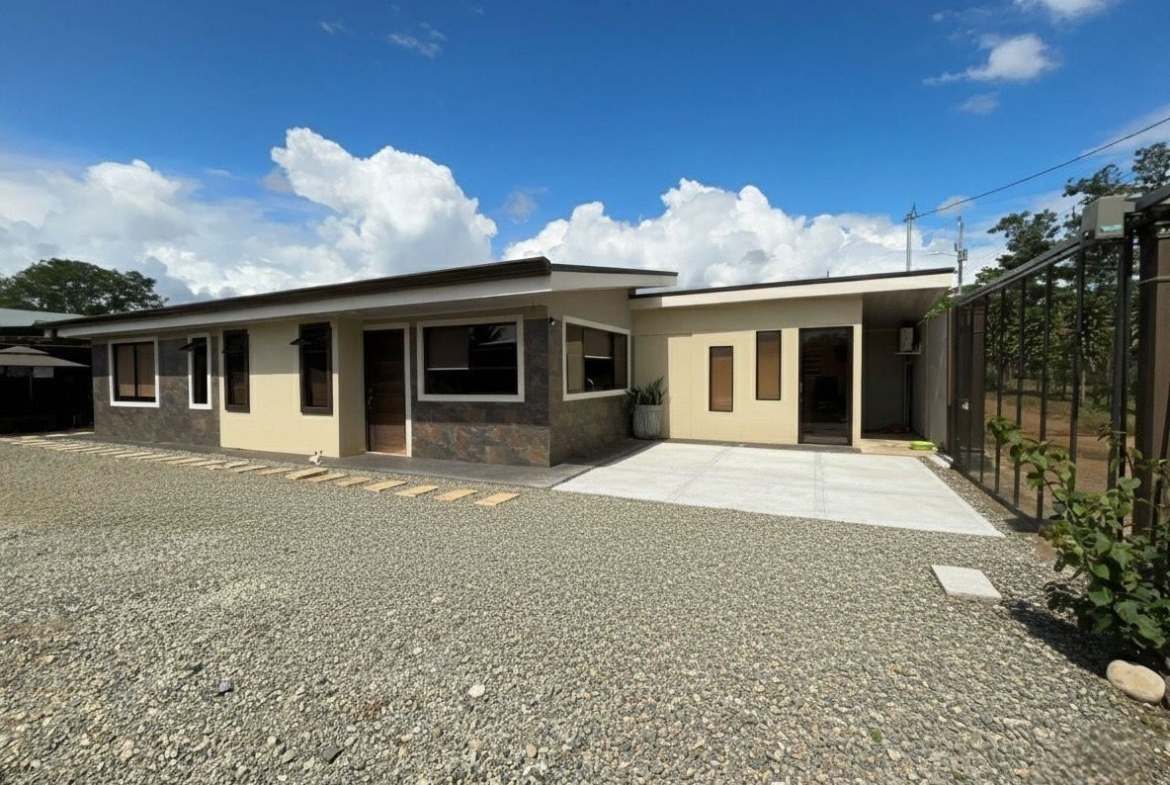 Front view of a modern, single-story house with beige walls, stone veneer accents, and a gravel yard under a blue sky.