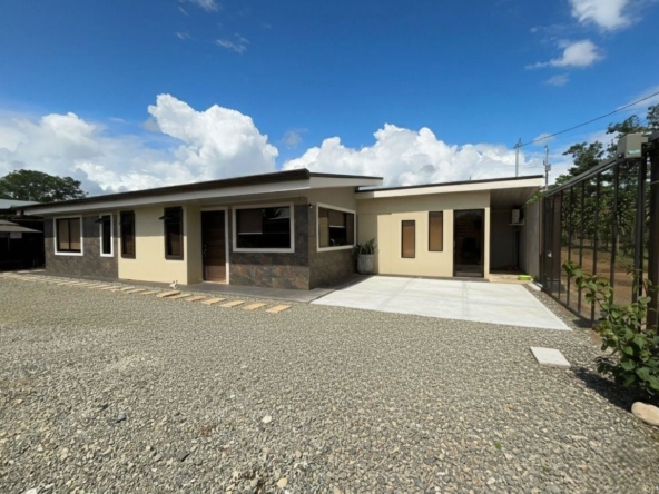 Front view of a modern, single-story house with beige walls, stone veneer accents, and a gravel yard under a blue sky.