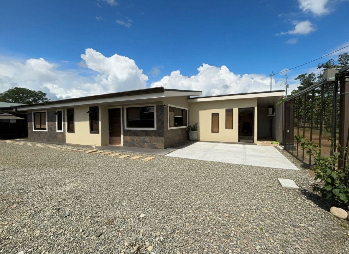 Front view of a modern, single-story house with beige walls, stone veneer accents, and a gravel yard under a blue sky.