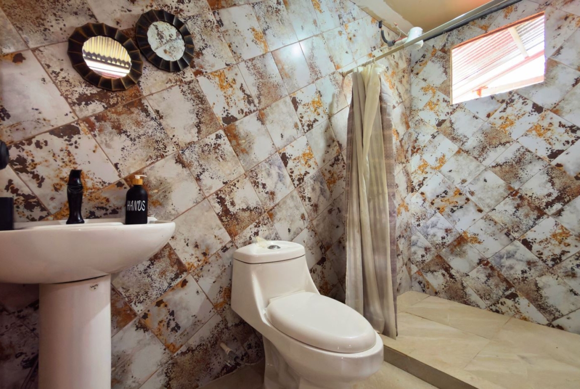 Bathroom with a white pedestal sink and toilet, wood- and rust-toned diagonally tiled walls, a shower area with a curtain and a small window above.