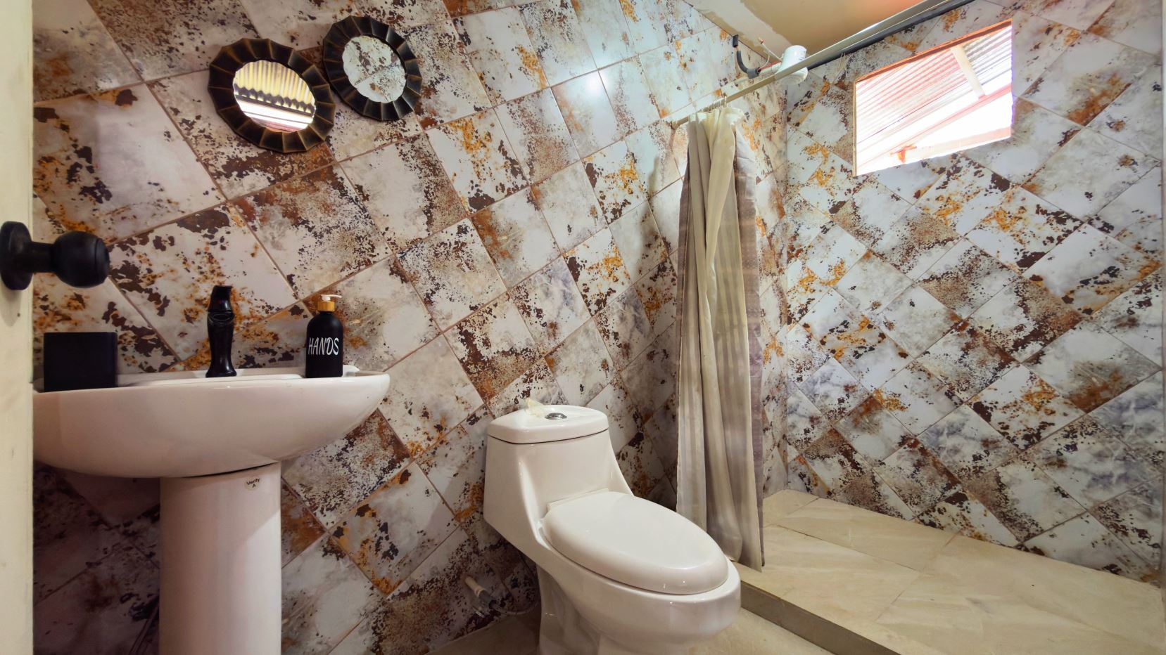 Bathroom with a white pedestal sink and toilet, wood- and rust-toned diagonally tiled walls, a shower area with a curtain and a small window above.