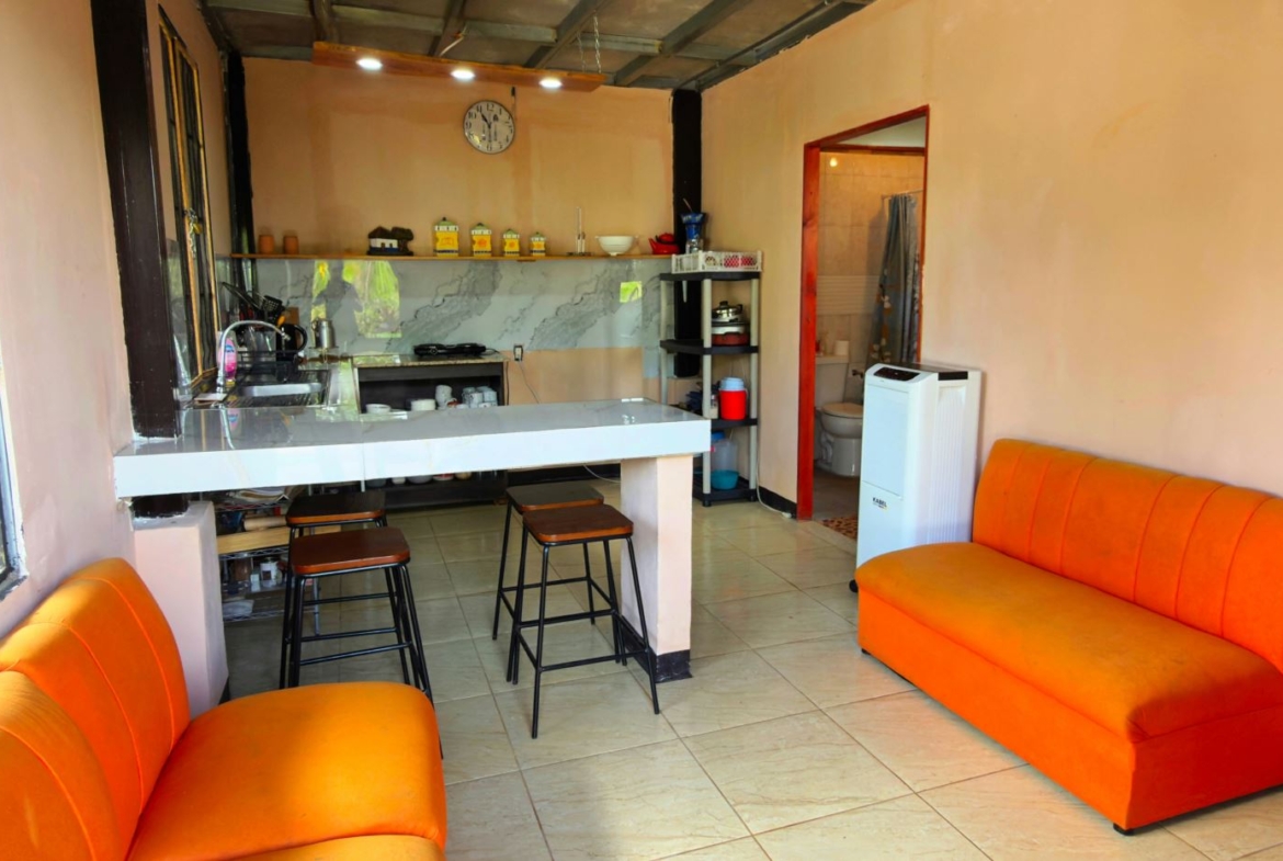 Cozy kitchenette and lounge with bright orange sofas, a white bar counter with two stools, marble backsplash, and a doorway to a bathroom.