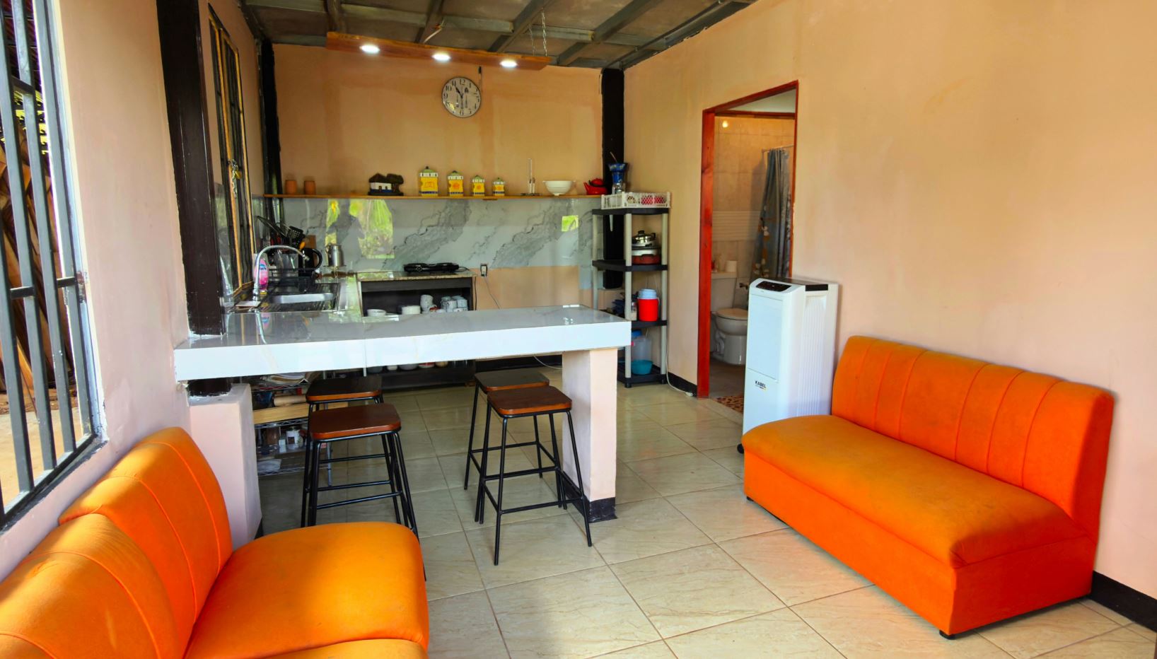 Cozy kitchenette and lounge with bright orange sofas, a white bar counter with two stools, marble backsplash, and a doorway to a bathroom.