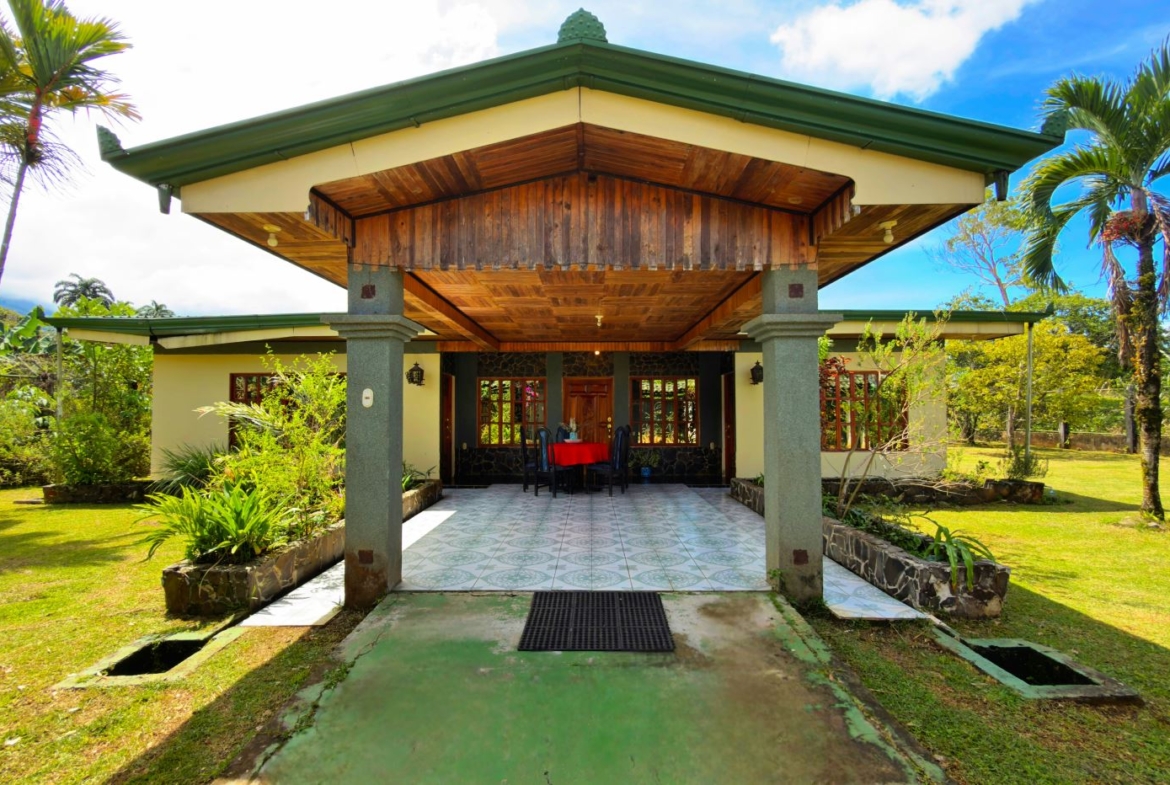 Open-air porch of a single-story tropical house with green roof and wooden ceiling, grey pillars, and a red-cloth dining table under the covered patio, surrounded by palm trees and a lawn.