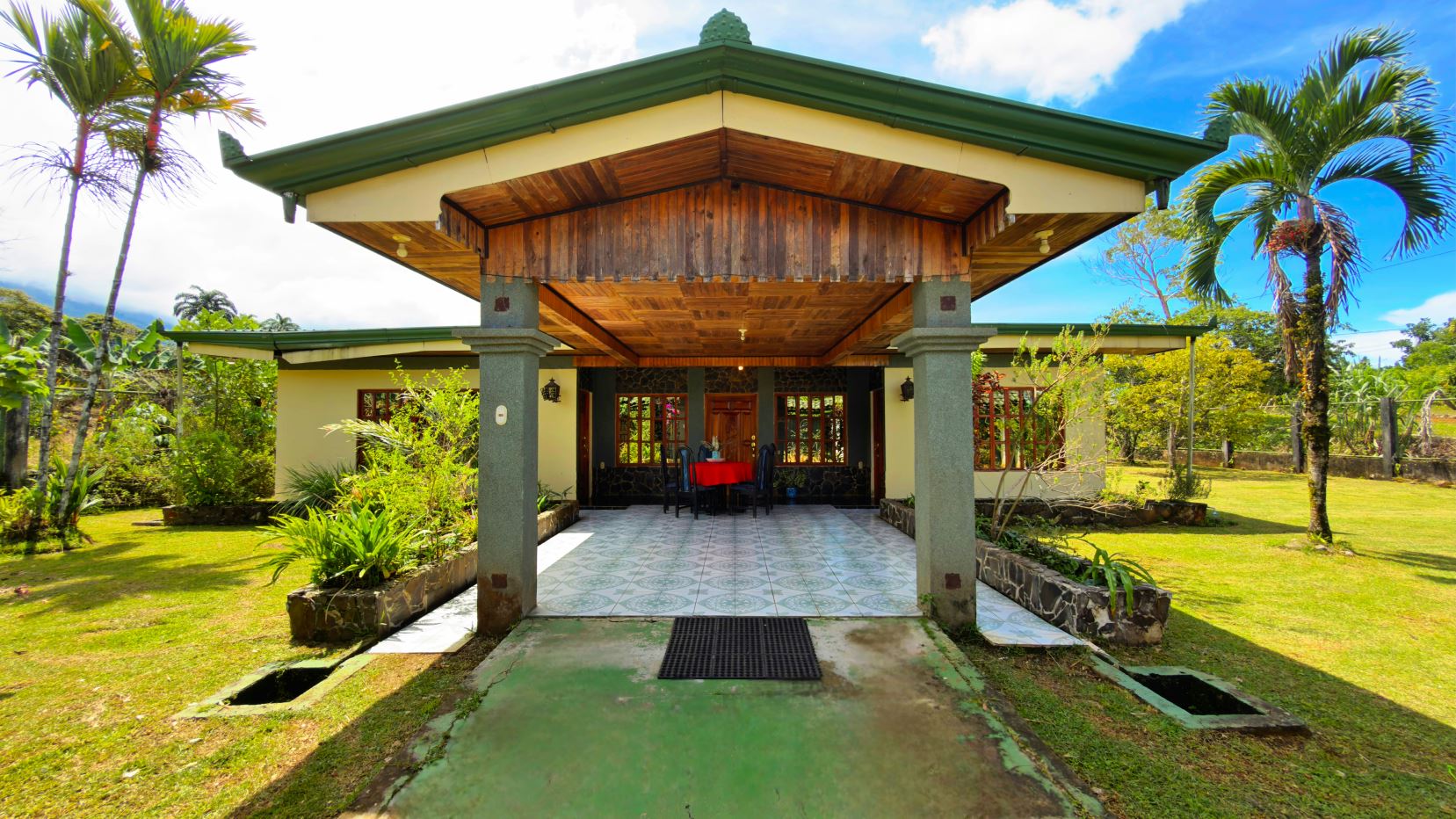 Open-air porch of a single-story tropical house with green roof and wooden ceiling, grey pillars, and a red-cloth dining table under the covered patio, surrounded by palm trees and a lawn.