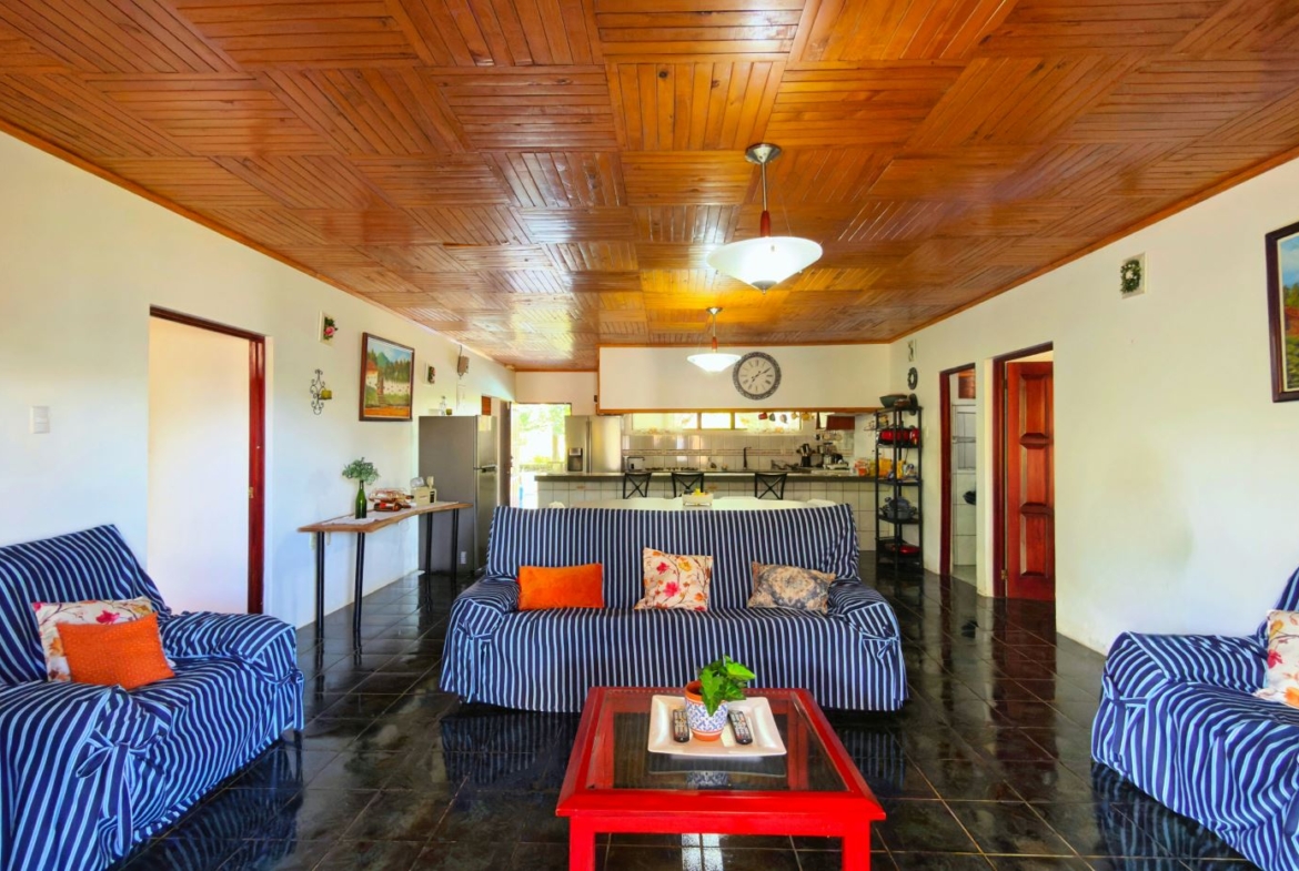 Living room with blue striped sofas and orange pillows, a red coffee table, and a kitchen area in the background.