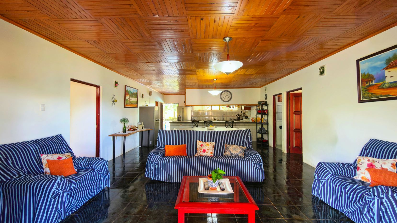 Living room with blue striped sofas and orange pillows, a red coffee table, and a kitchen area in the background.