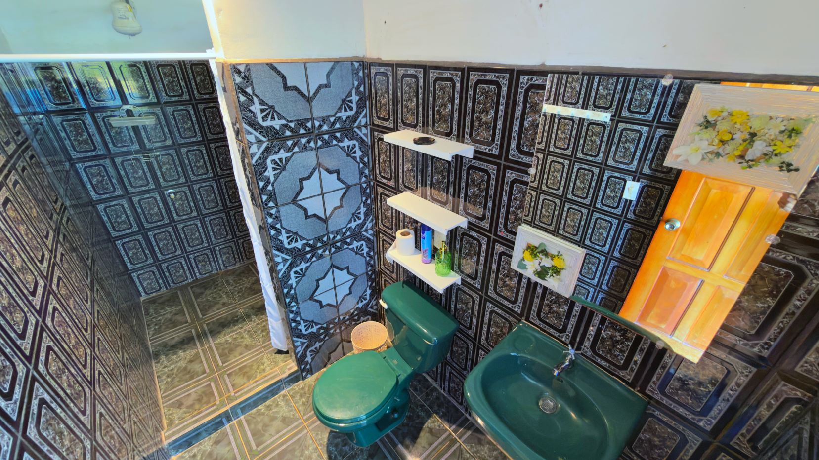 Bathroom with bold black geometric tiles, green toilet and sink, glass shower, and white shelves holding toiletries and paper towels.
