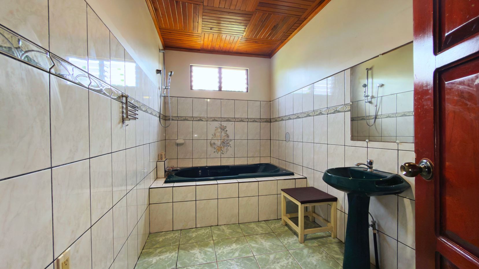 Bathroom with white tiled walls, a green built-in tub, a matching green pedestal sink, and a wooden ceiling; doorway visible on the right side.