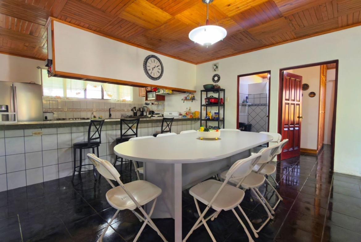 Open kitchen and dining area with a long white table surrounded by white folding chairs, dark tile floor, and a wood-paneled ceiling.