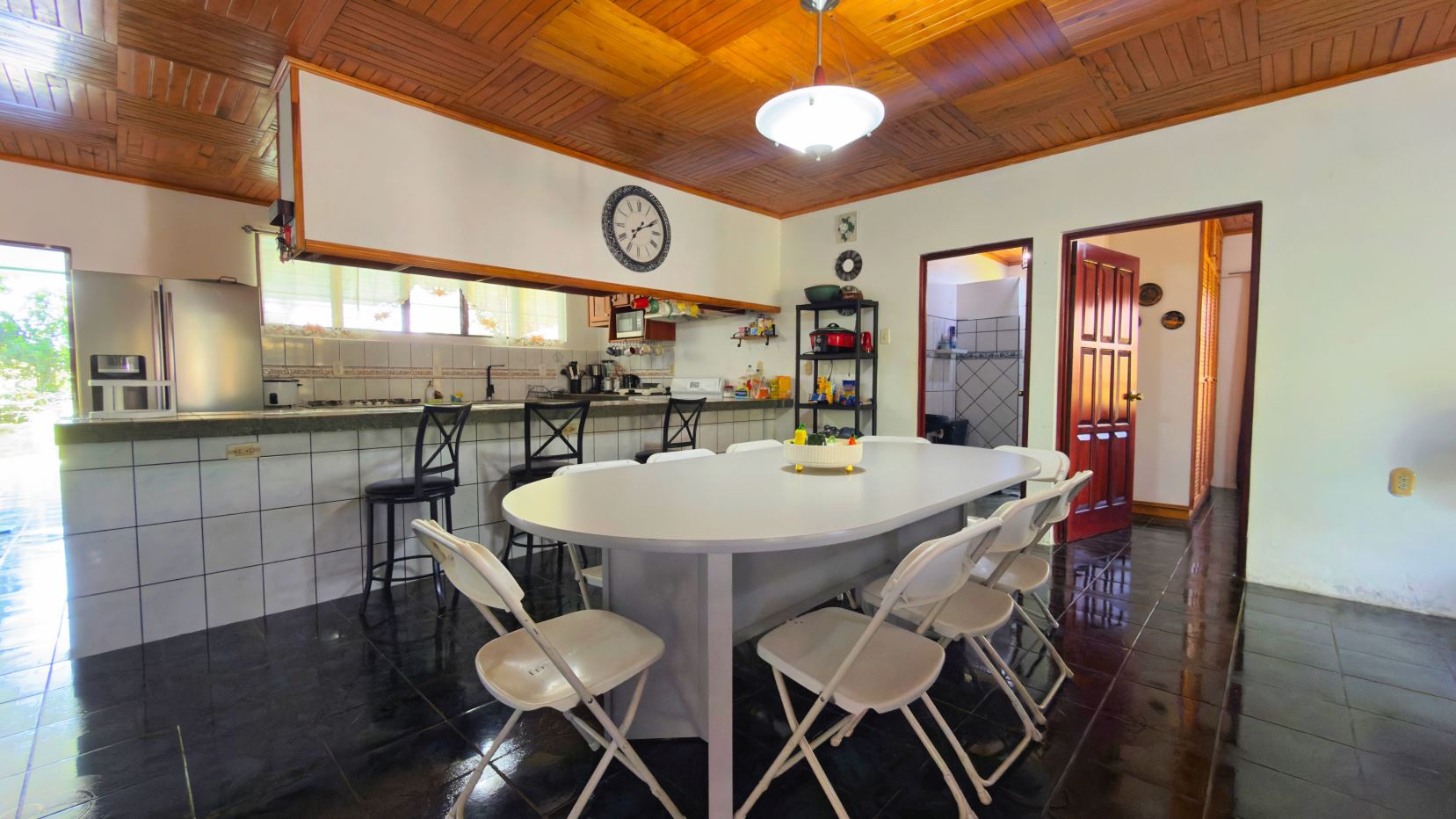 Open kitchen and dining area with a long white table surrounded by white folding chairs, dark tile floor, and a wood-paneled ceiling.