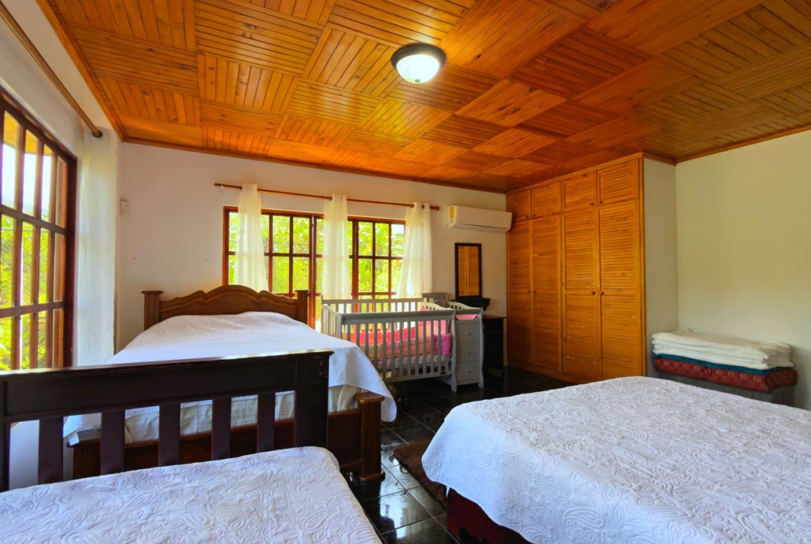 Bright bedroom with two large beds, a baby crib, and a wooden wardrobe along the wall. Large windows let in natural light.