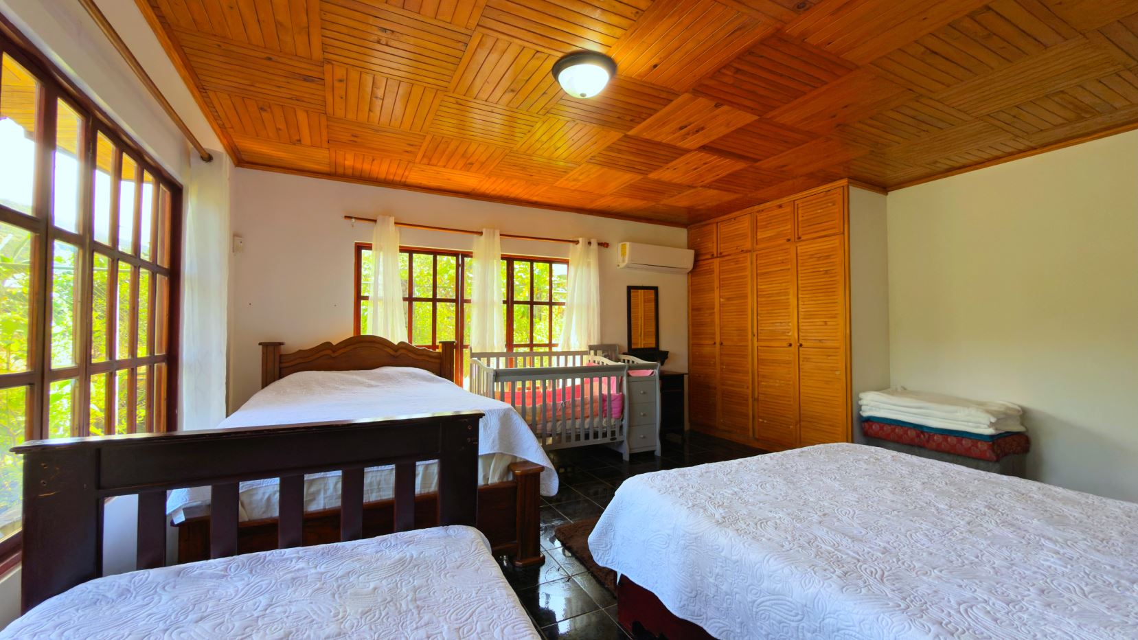 Bright bedroom with two large beds, a baby crib, and a wooden wardrobe along the wall. Large windows let in natural light.