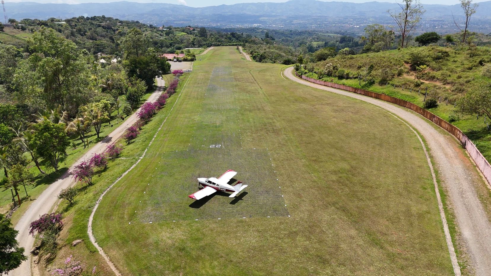 Small red-and-white single-engine plane on a grassy airstrip, with runway markers and surrounding trees beyond.