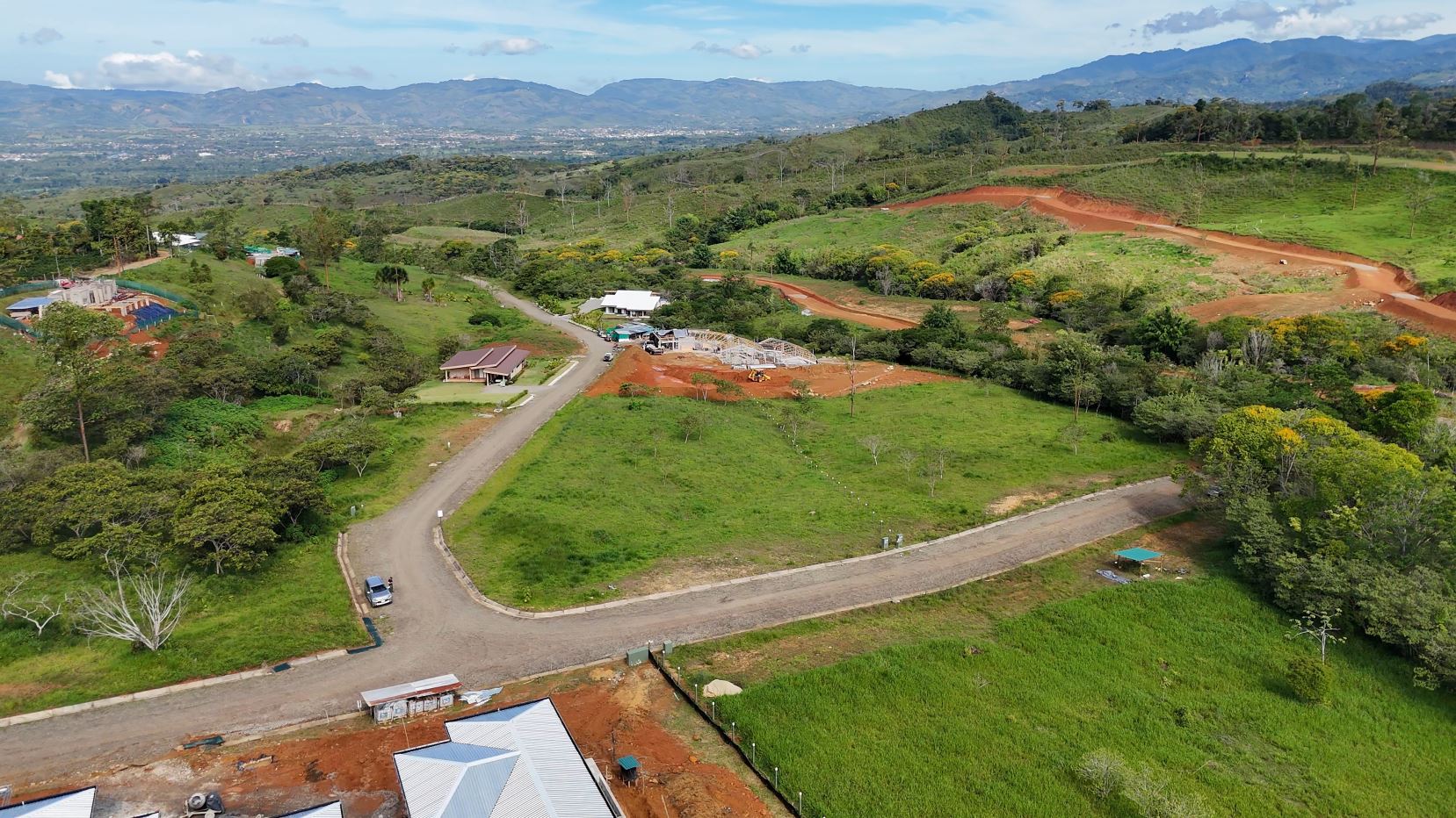 Aerial view of a rural landscape with winding dirt roads, building sites, green fields, and distant mountains under a blue sky.