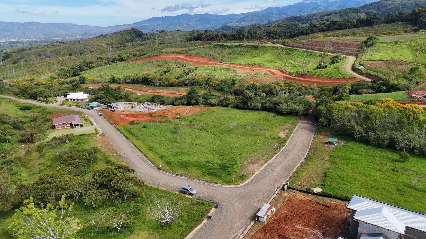 Aerial view of a countryside with winding roads, green fields, and a construction site on red soil near houses.
