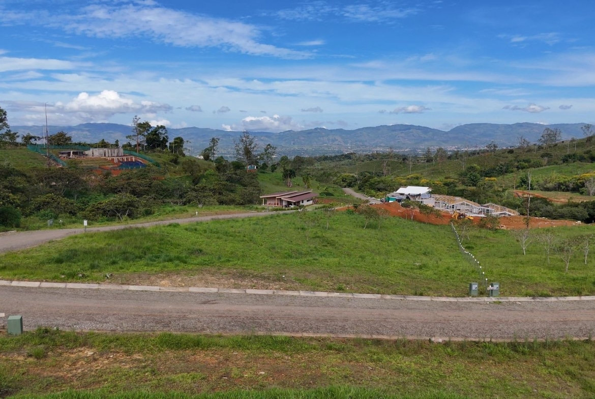 Rural hillside with a winding road, green fields, and a building under construction in the distance against mountains and a blue sky
