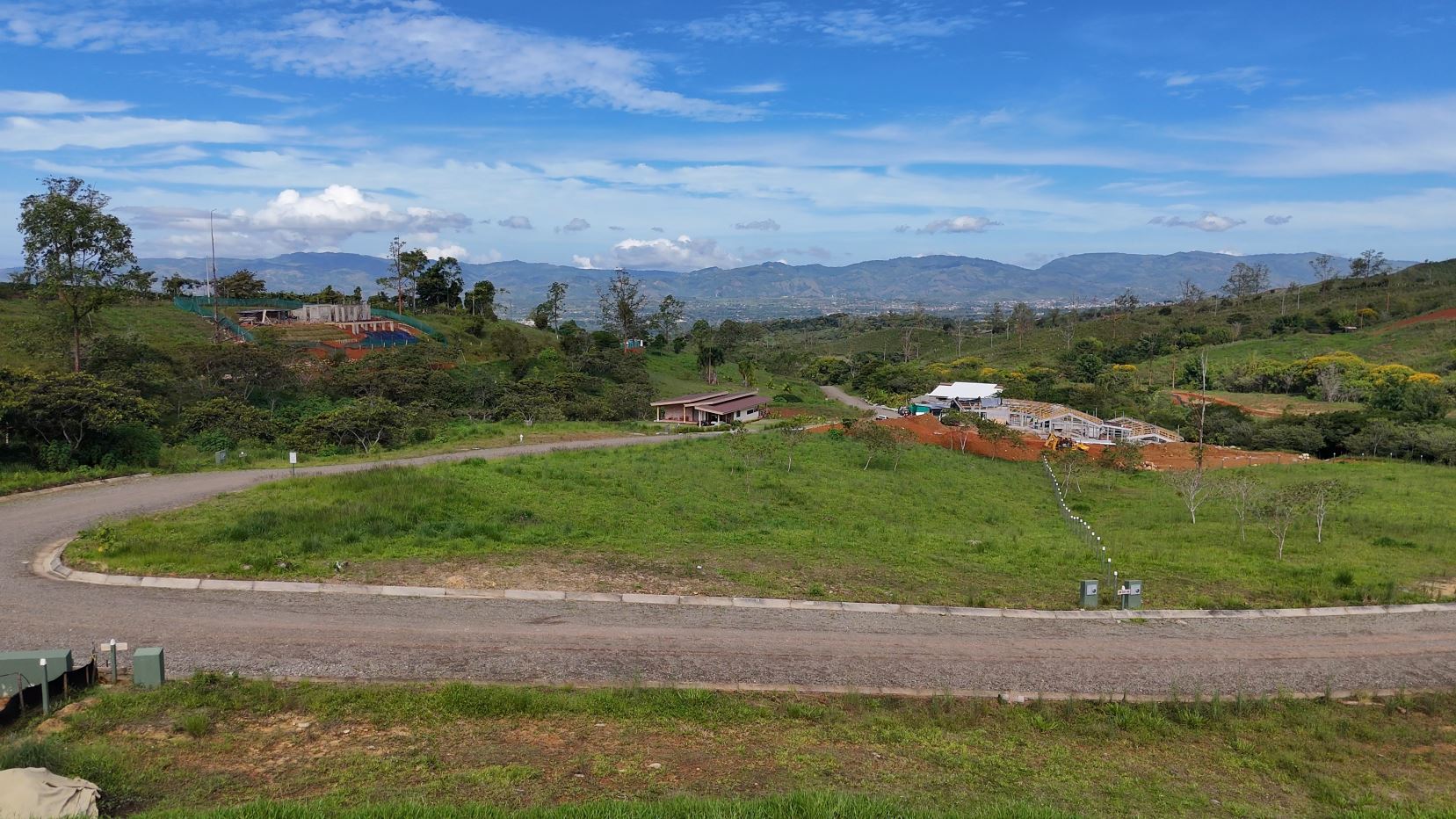 Rural hillside with a winding road, green fields, and a building under construction in the distance against mountains and a blue sky