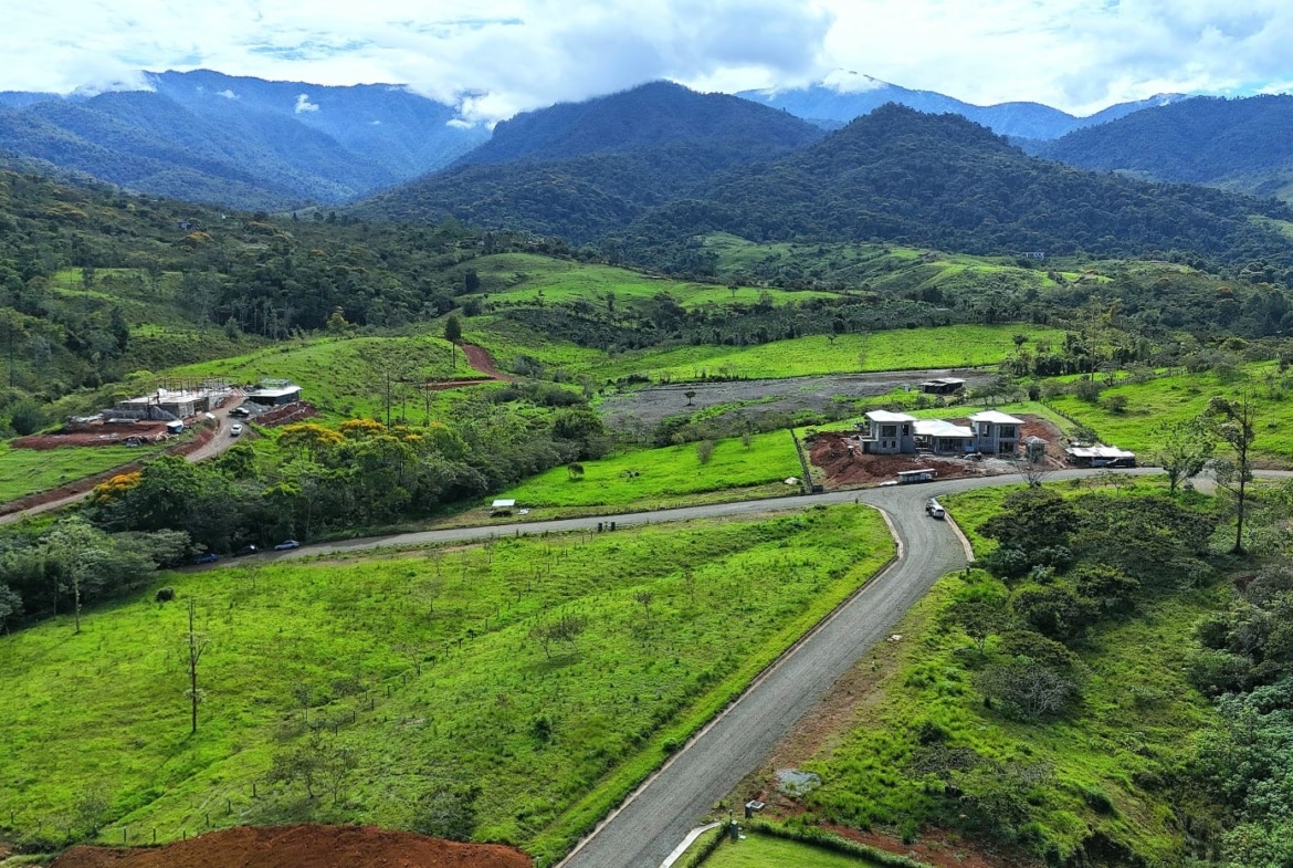 Rural hillside with green fields, a winding road, and a small construction site with two buildings in the midground, mountains in the distance.