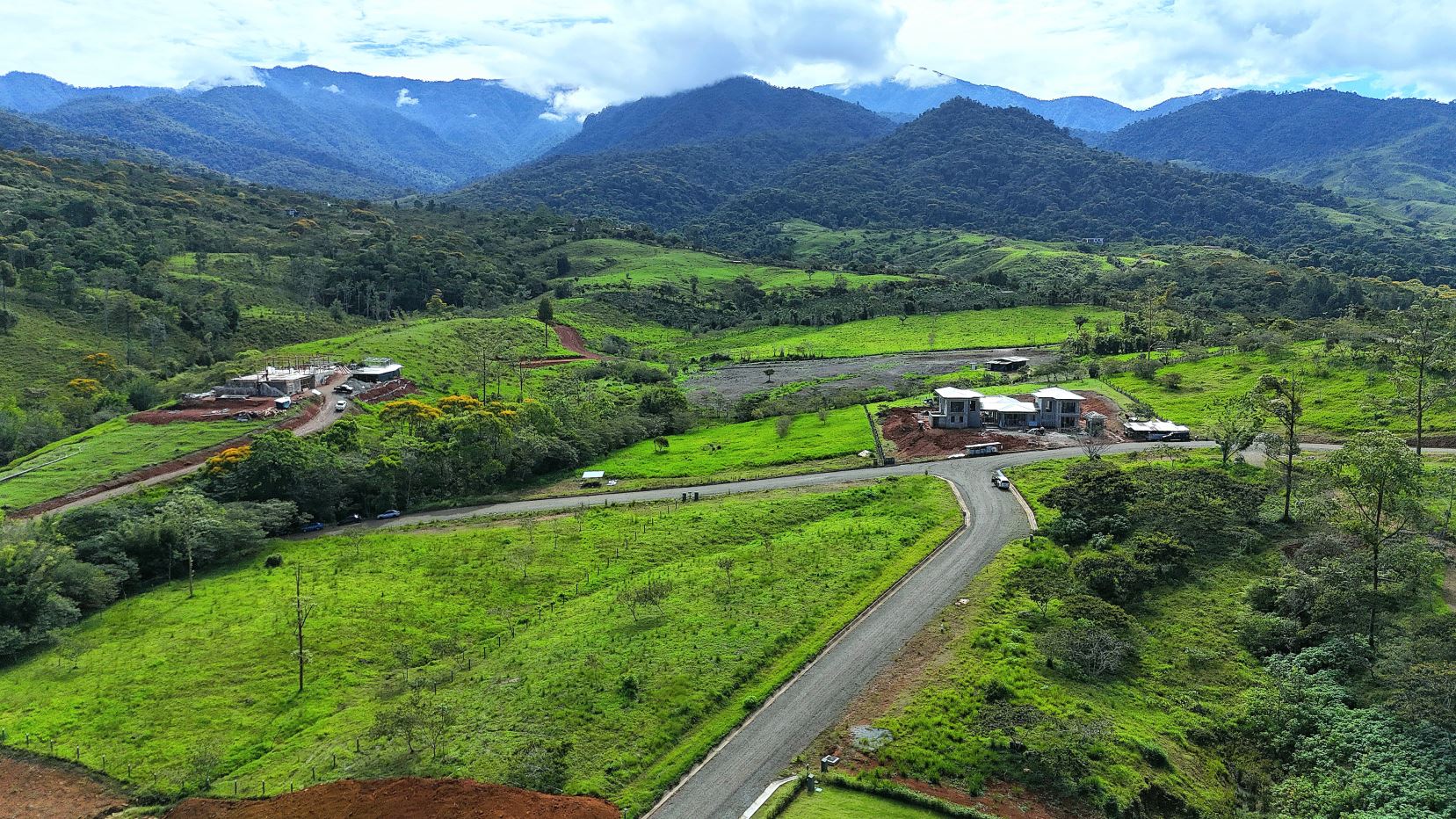 Rural hillside with green fields, a winding road, and a small construction site with two buildings in the midground, mountains in the distance.