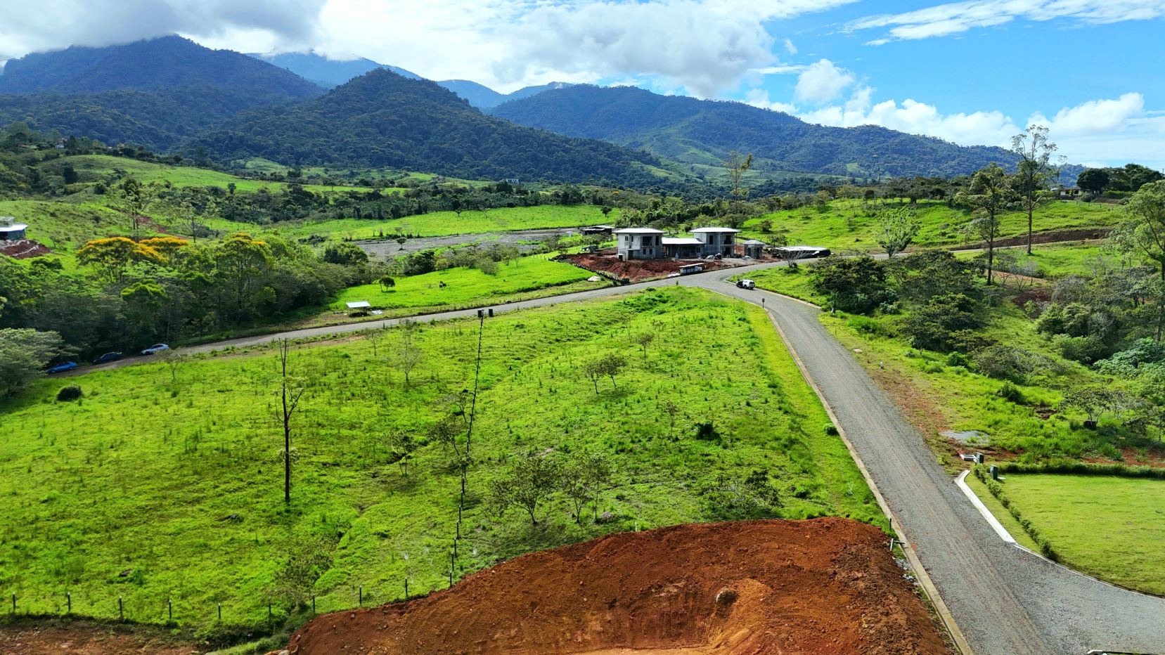 Lush green valley with rolling hills and distant mountains; a curved road leads to a small construction site with white buildings at a crossroads.