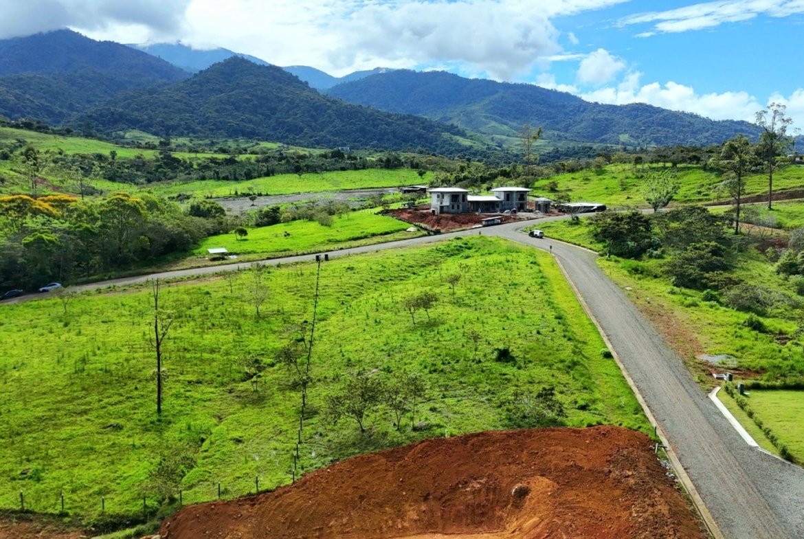 Green valley with a winding road, a small construction site, and distant hills under a blue sky with clouds.