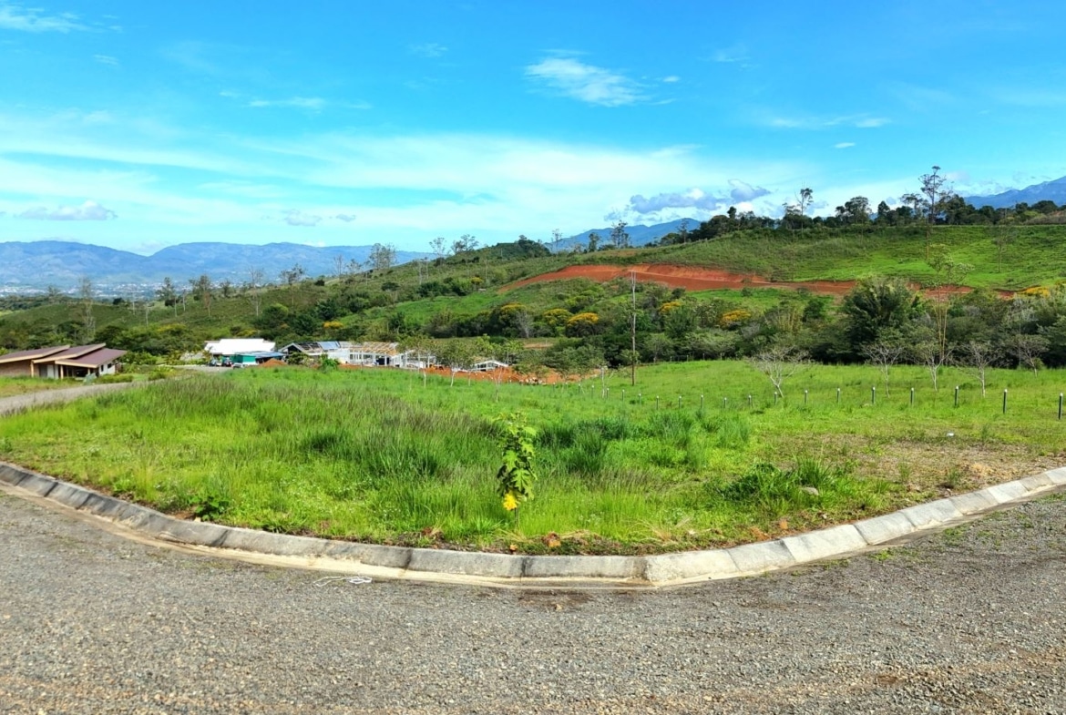 Curved road framing a grassy plot in a rural hillside landscape with distant mountains and blue sky widening above.