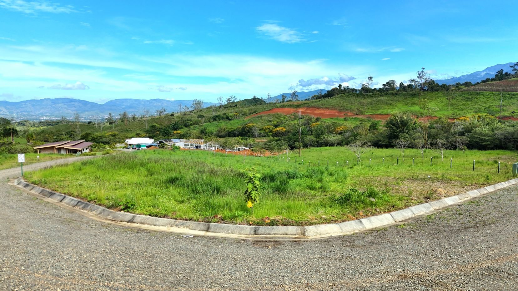 Curved road framing a grassy plot in a rural hillside landscape with distant mountains and blue sky widening above.