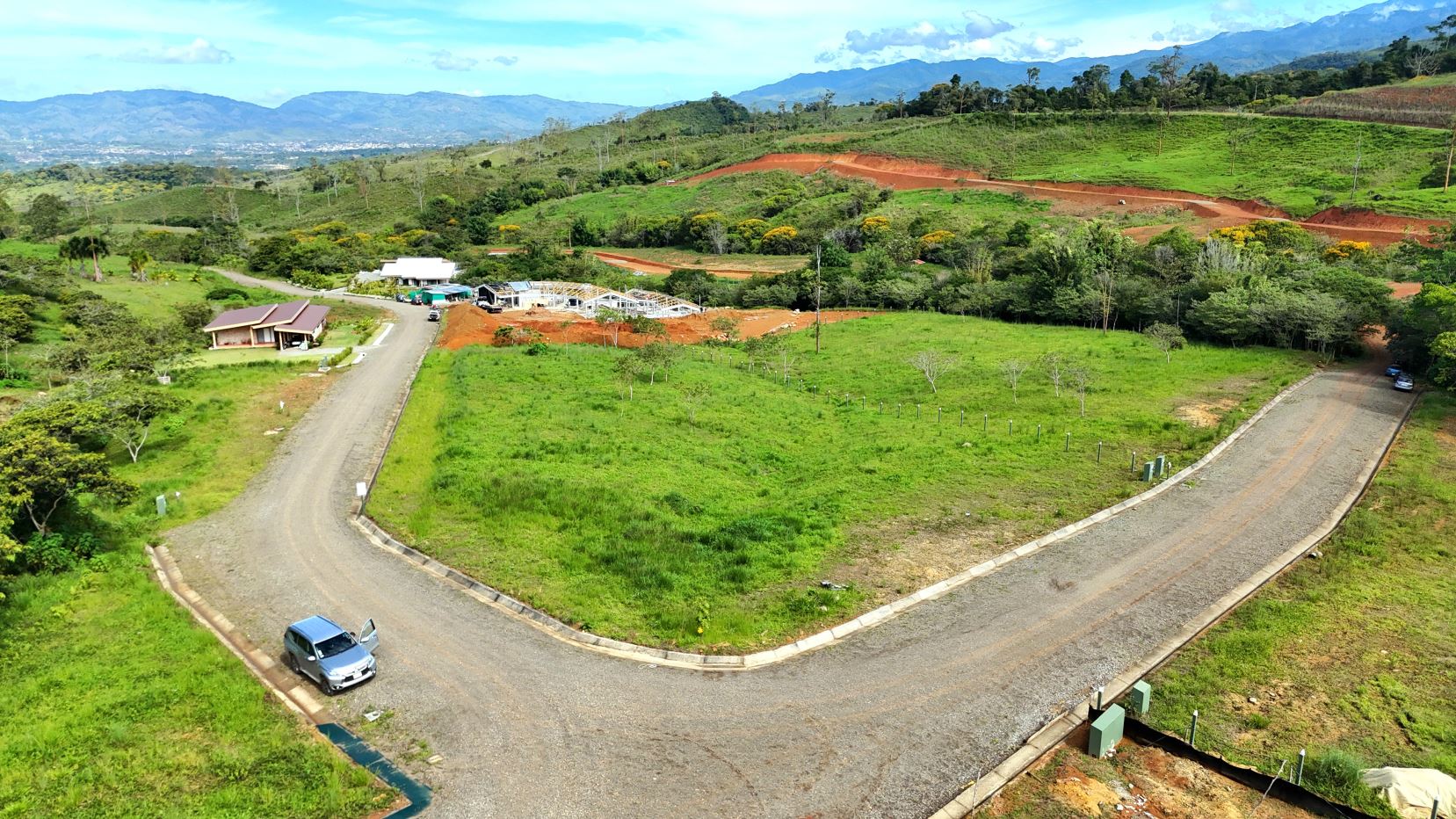 Aerial view of a curved gravel road looping around a grassy lot with a construction site and small buildings in a rural valley.