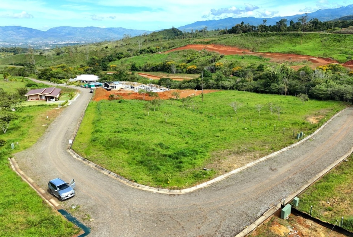 Aerial view of a curved gravel road looping through a green development site with a small building and construction area, hills in the distance.