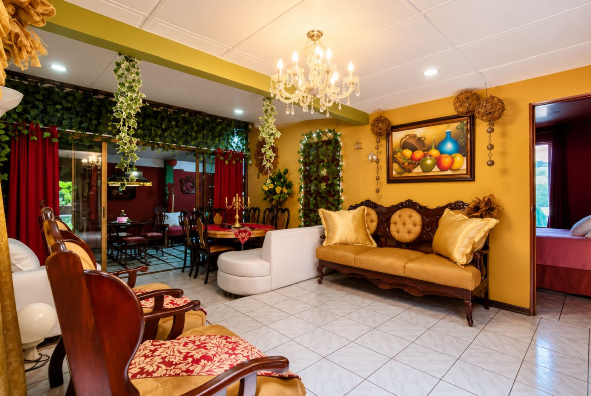 Elegant living room with yellow walls, chandelier, and tufted sofa; potted plants and framed fruit still-life art on wall.