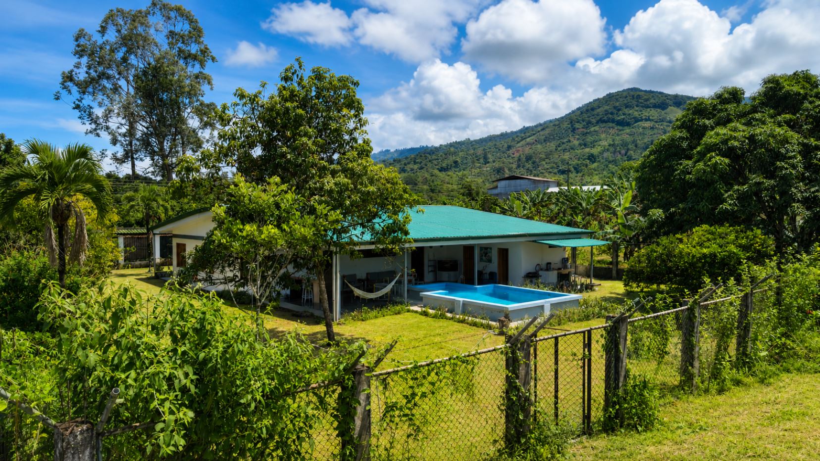 Single-story white house with a turquoise roof, hammock in the yard, and a small rectangular pool, surrounded by lush tropical trees and a chain-link fence, with hills in the distance.