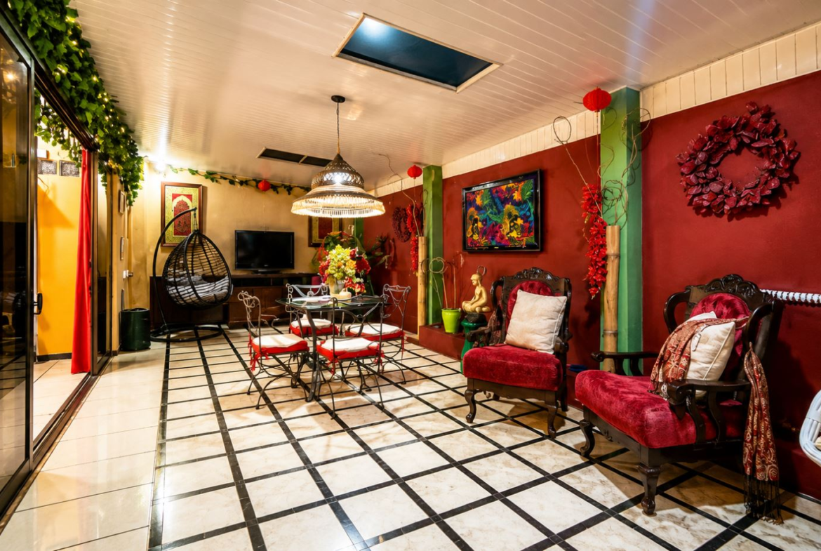Colorful indoor lounge with red walls, ornate chairs, a glass dining table, and a hanging egg chair surrounded by greenery.