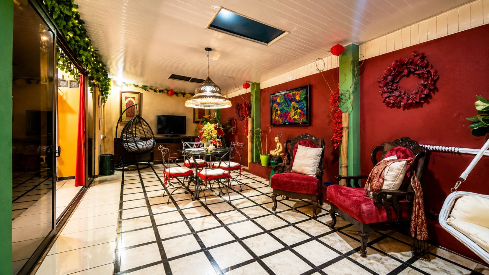 Colorful indoor lounge with red walls, ornate chairs, a glass dining table, and a hanging egg chair surrounded by greenery.