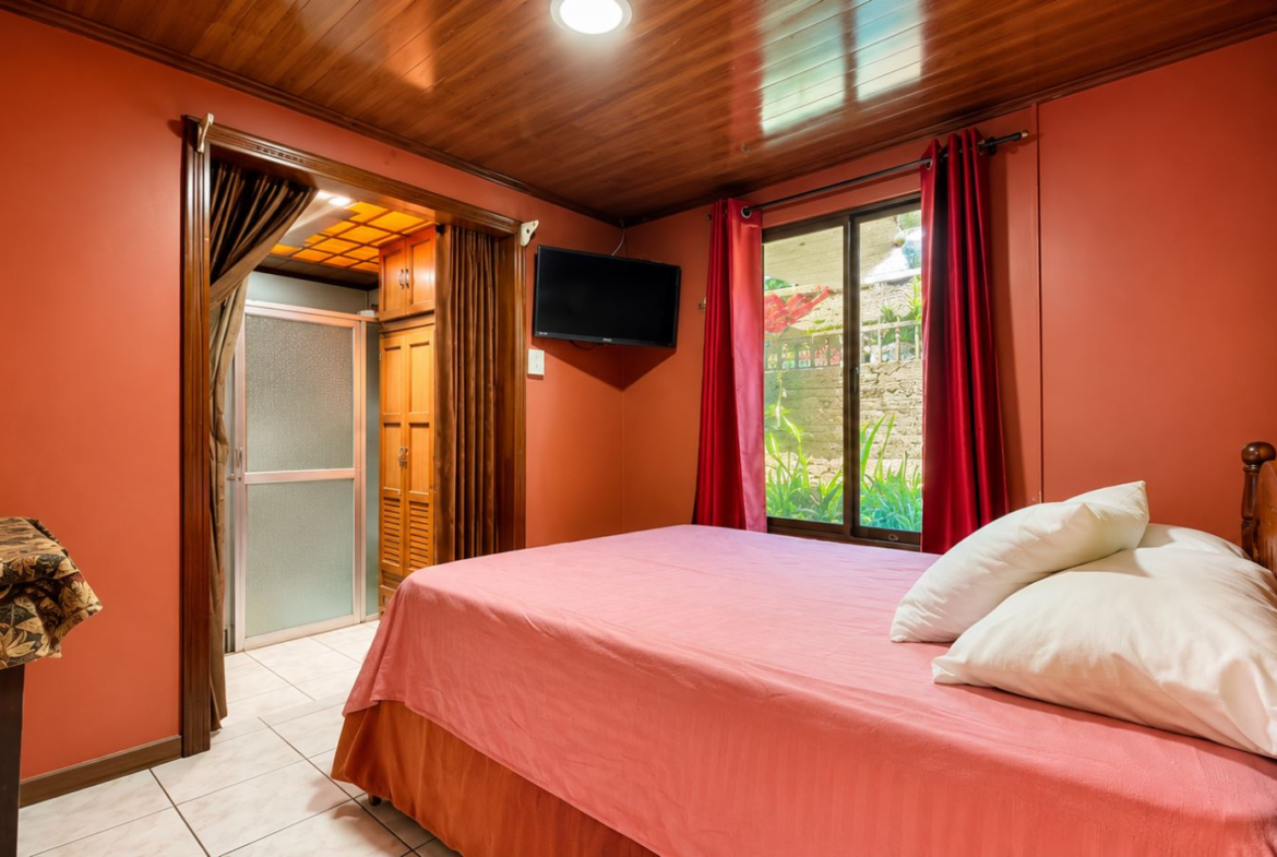 Bedroom with coral walls, a pink bedspread, white pillows, a wall-mounted TV, and a window with red curtains.